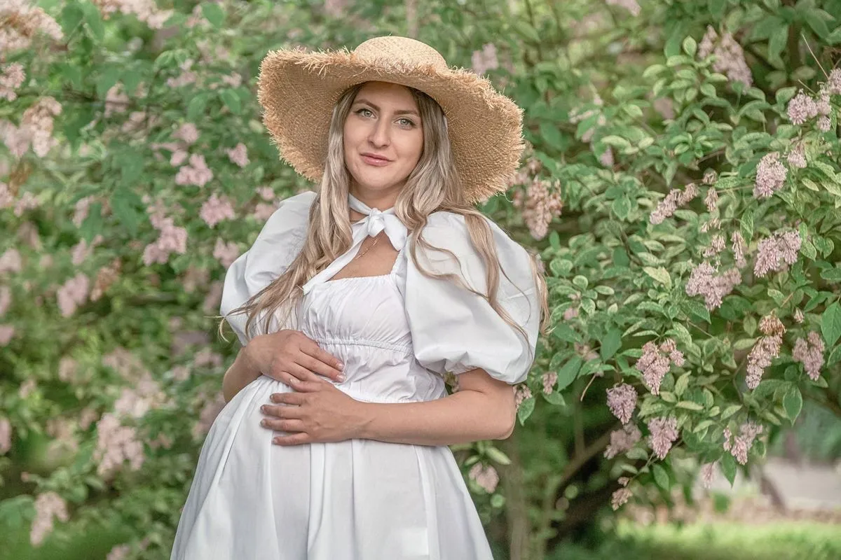 Gestante de chapéu de palha e vestido branco, com as mãos na barriga, ao lado de arbustos floridos